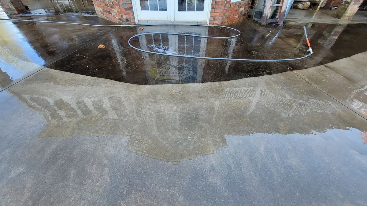 A concrete patio outside a pair of French doors, dark with grime near the doors and clean in front, with a hose lying on the wet surface